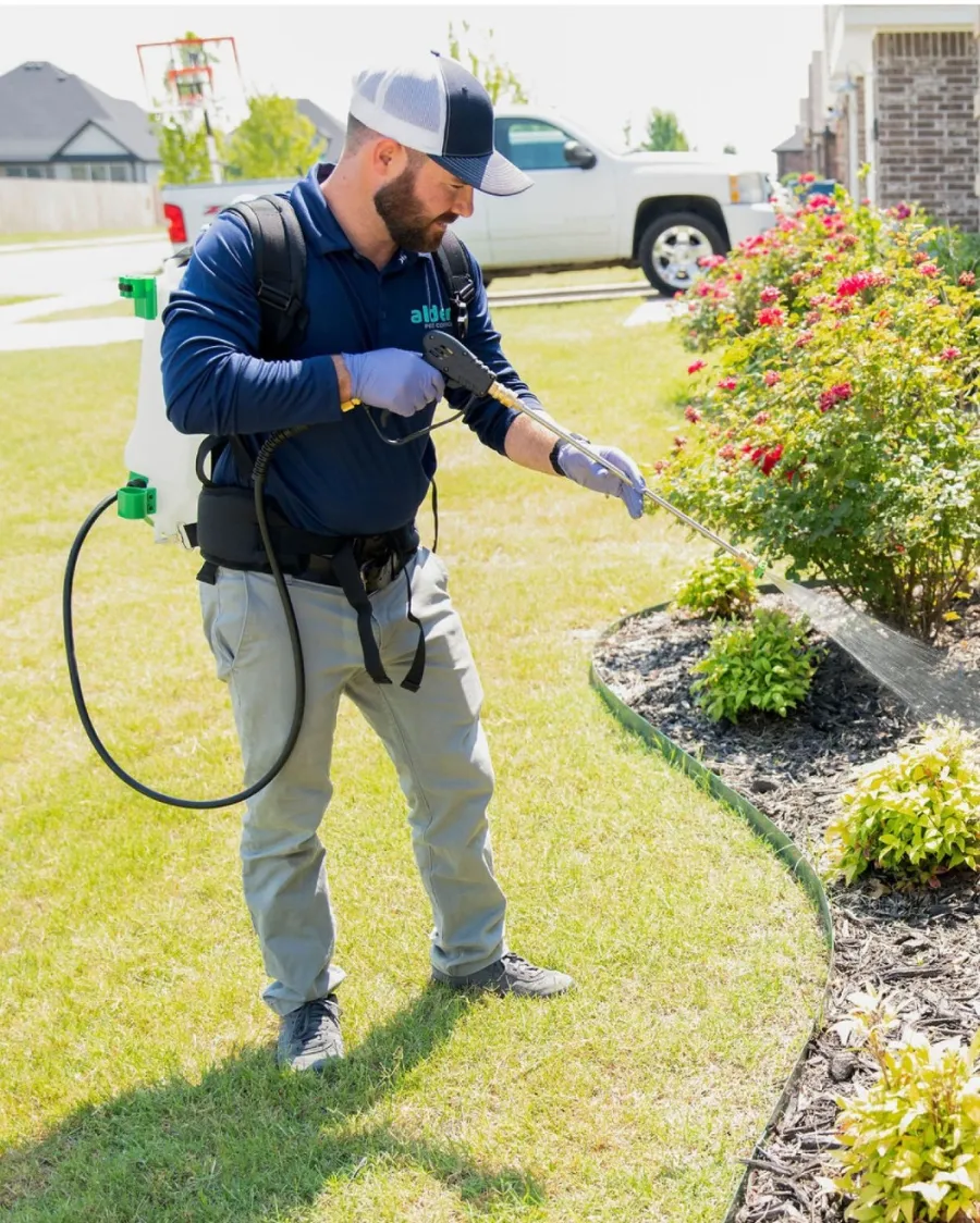 A professional applying pesticide to a home garden with lush greenery and flowers.
