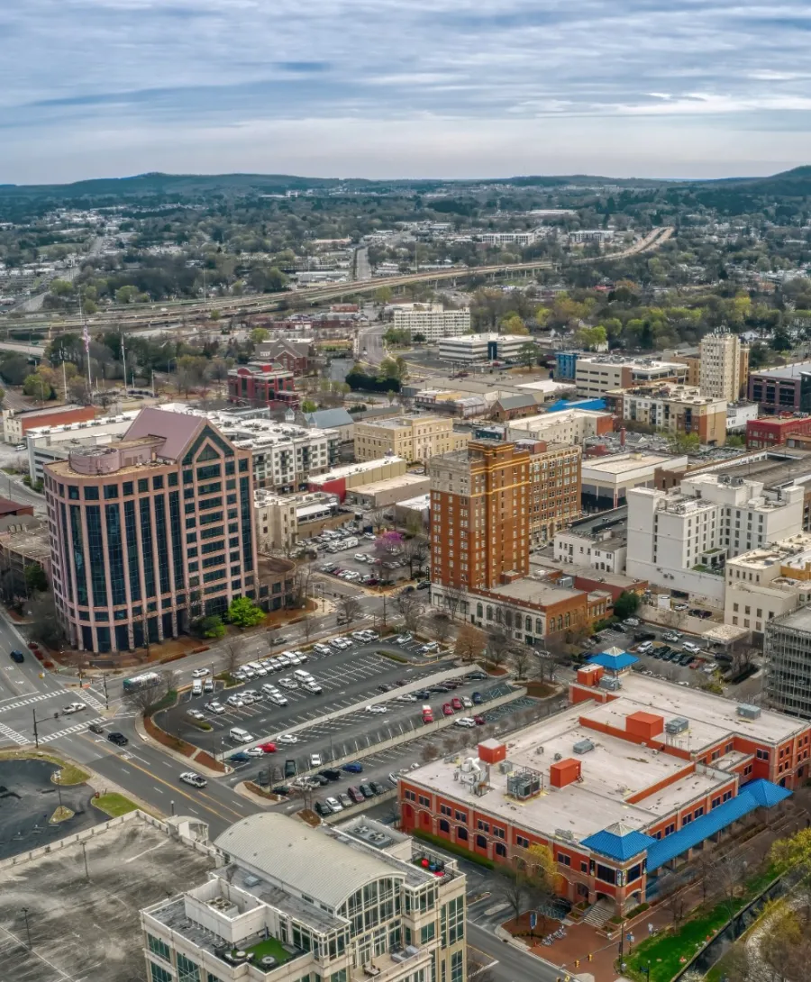 Aerial view of a cityscape with commercial buildings, streets, green park, and surrounding hills under a cloudy sky.