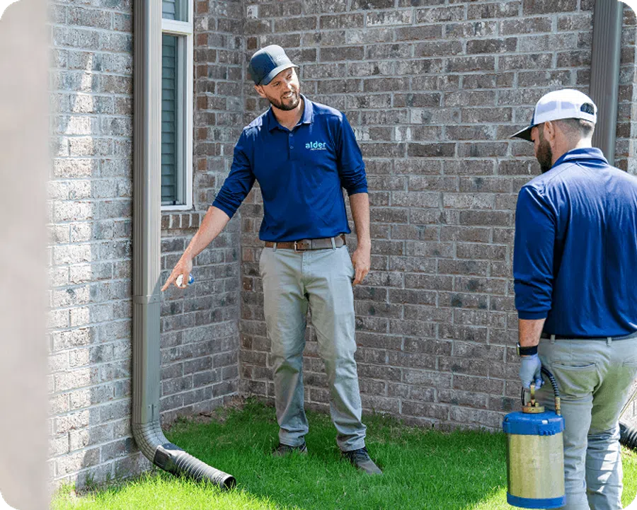 Two pest control workers inspecting a home's exterior brick wall and downspout on green grass