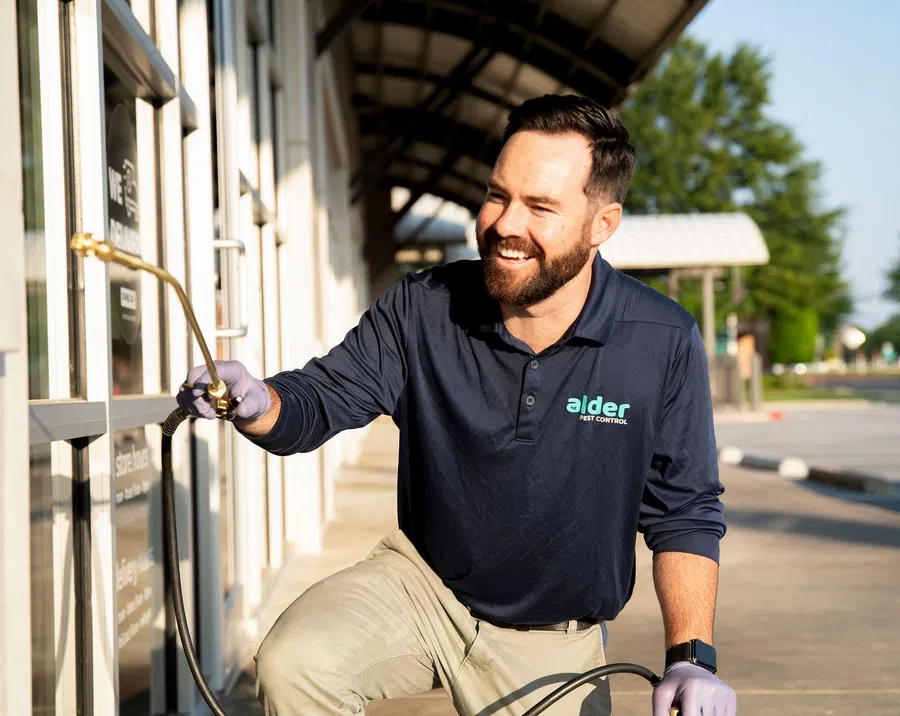 Smiling pest control technician in dark shirt spraying insecticide on a commercial building exterior window.