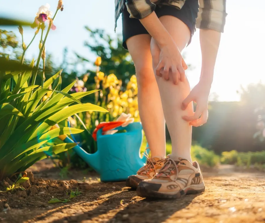 kid outdoors in garden touching leg, wearing hiking shoes with watering can and plants nearby in sunlight.