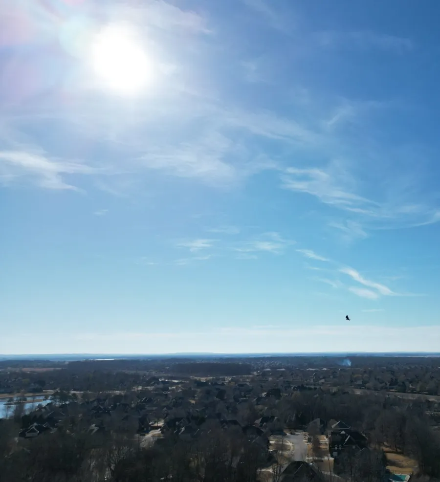 Clear blue sky with the sun shining over a suburban area featuring trees, houses, and small water bodies under a sunny day.