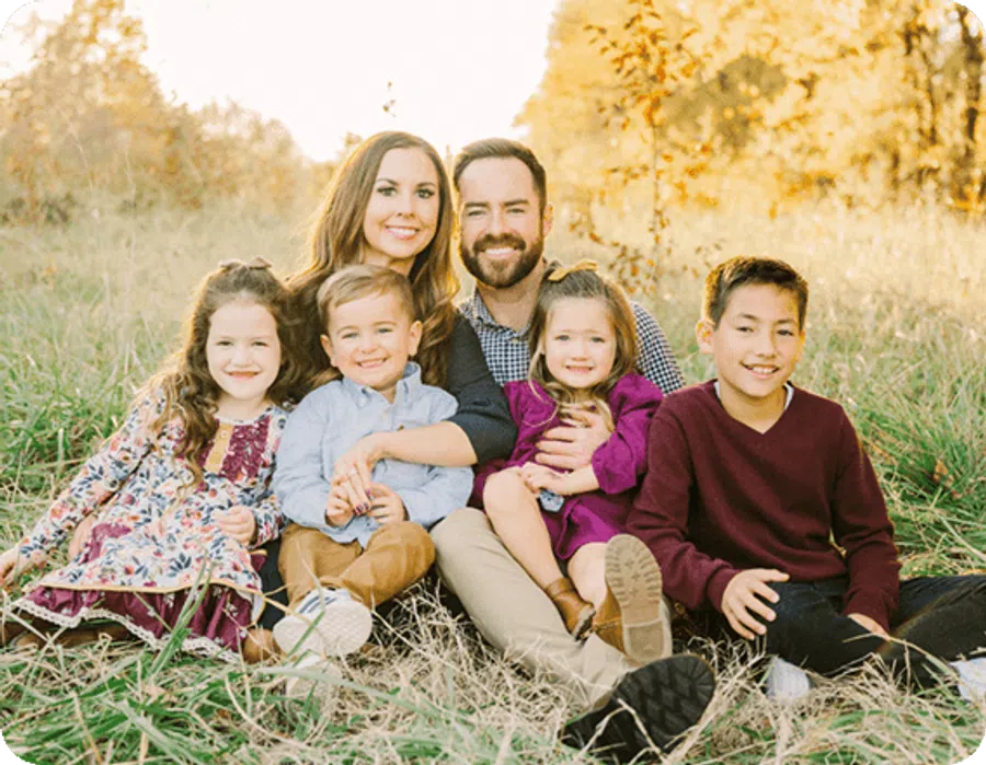 Smiling family of six sitting together in a grassy field during golden hour with warm sunlight and autumn trees.