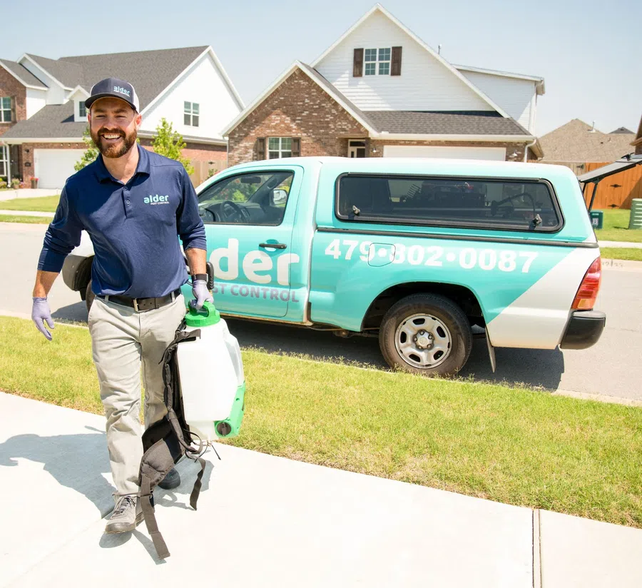 Smiling pest control technician walking on sidewalk with equipment near branded company pickup truck in suburban neighborhood