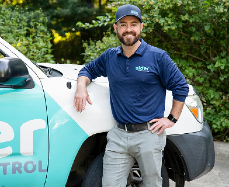 Alder pest control technician smiles beside a service vehicle in a green landscape.