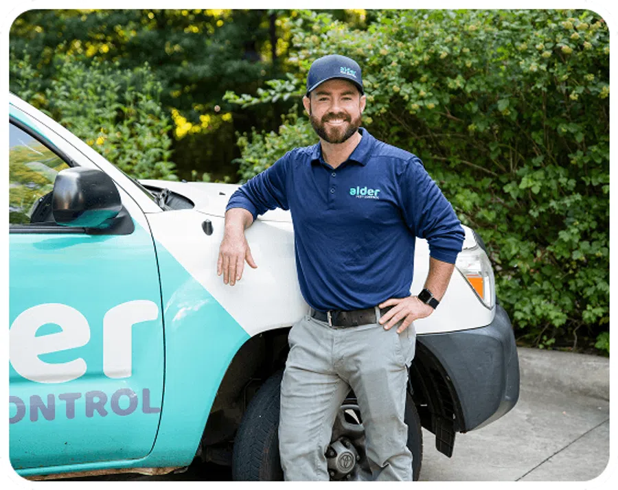 Smiling pest control technician in company uniform leaning on branded service truck outdoors with greenery background