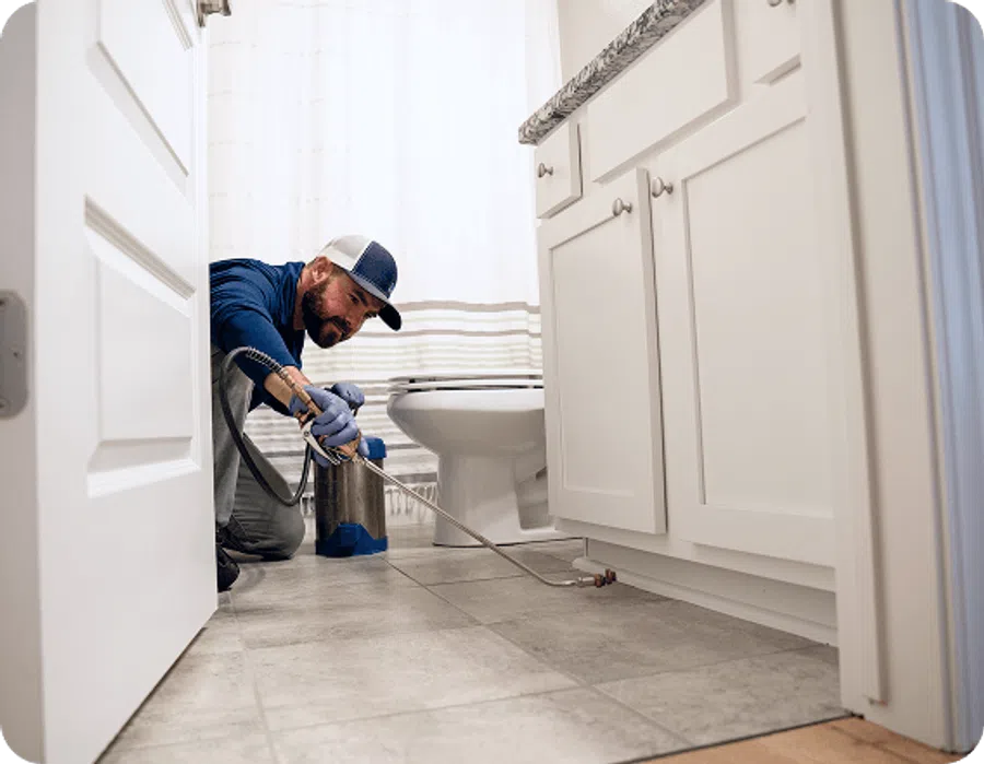 Pest control technician spraying insecticide along bathroom cabinet baseboards in a home bathroom.