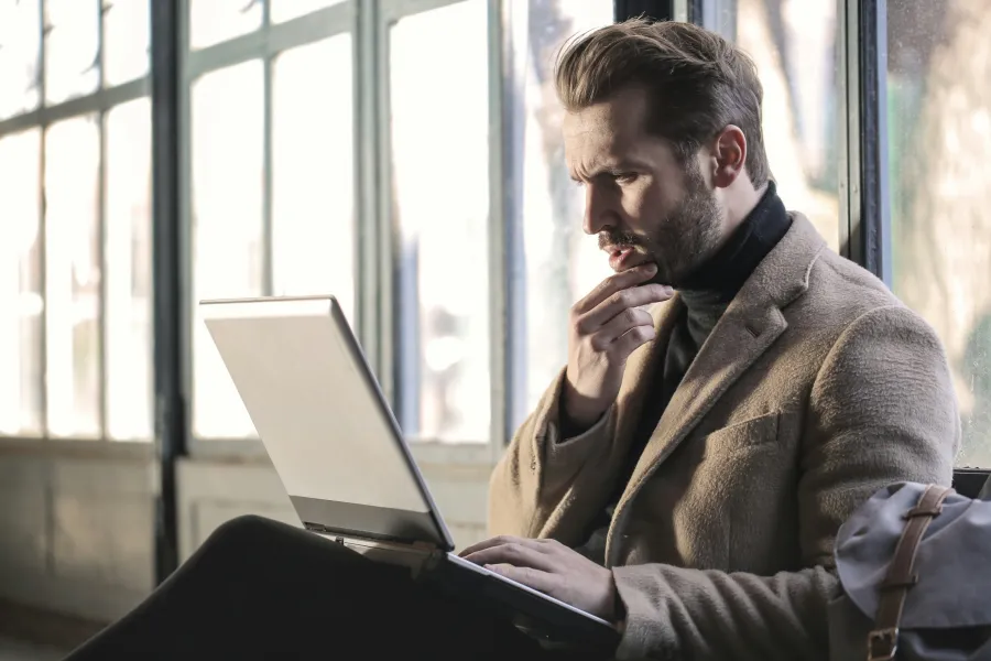 Young man in beige coat working thoughtfully on a laptop by a large window with natural light.