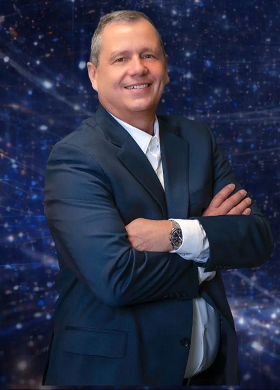 Smiling middle-aged man in a navy suit with arms crossed against a blue digital network background.