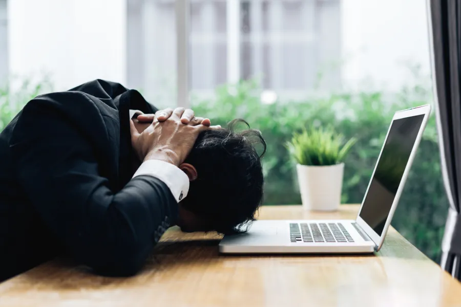 Frustrated businessman in suit with head on hands at desk next to open laptop and potted plant