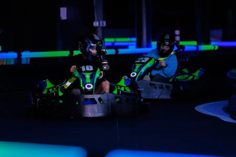 Two people racing go-karts indoors with neon green and blue lighting in a dark setting.