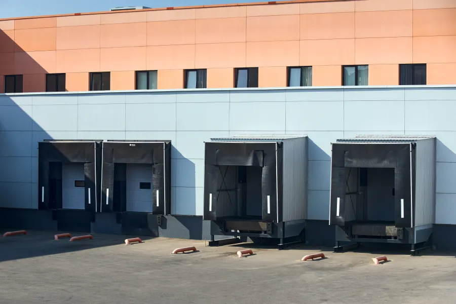 Four loading dock bays with black seals on a warehouse exterior beneath orange and blue walls under clear sunlight