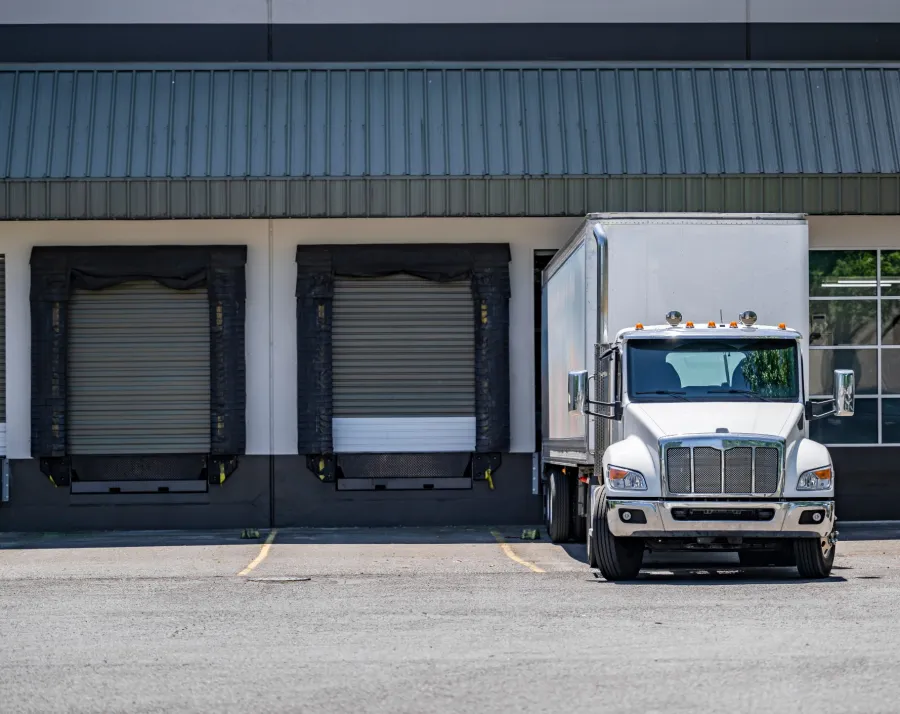 White semi-truck parked in front of a loading dock at a warehouse building under bright daylight.