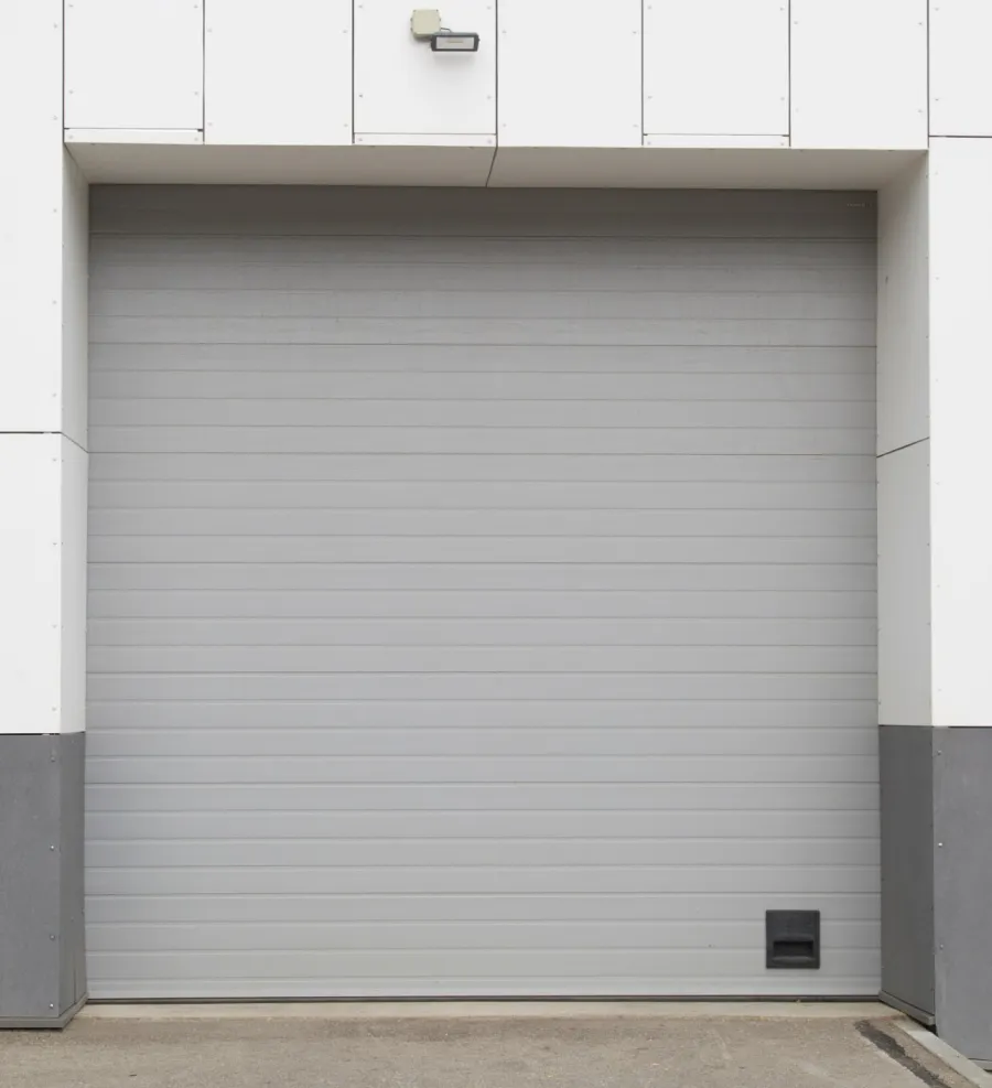 Closed gray industrial garage door with white and gray panel building exterior and security light above.
