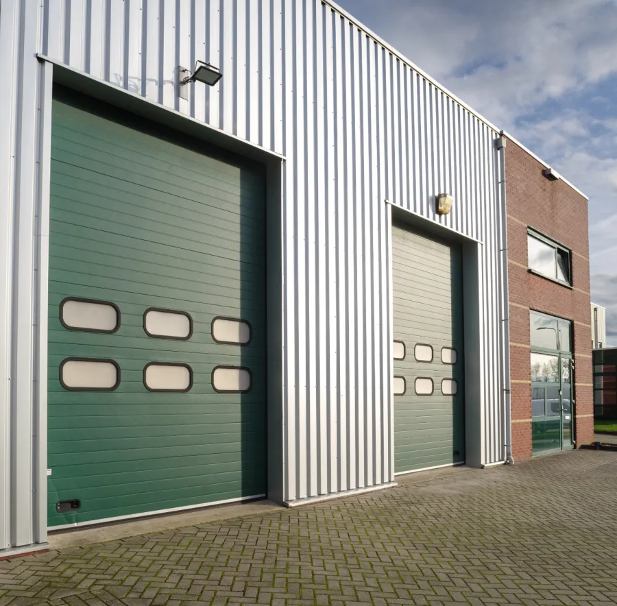 Industrial building with two green sectional overhead garage doors and brick and metal exterior under a partly cloudy sky