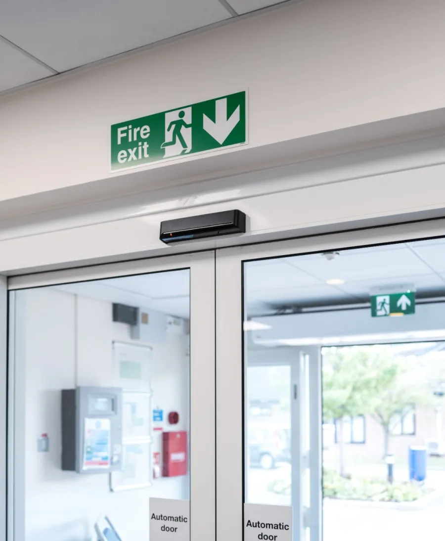 Automatic glass fire exit doors in a building with a green fire exit sign overhead and outdoor view visible.
