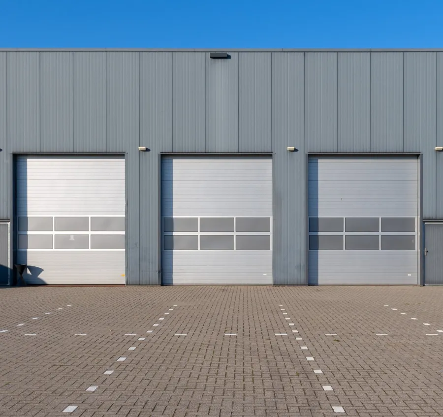 Empty parking lot with marked spaces in front of four closed industrial warehouse doors under clear blue sky