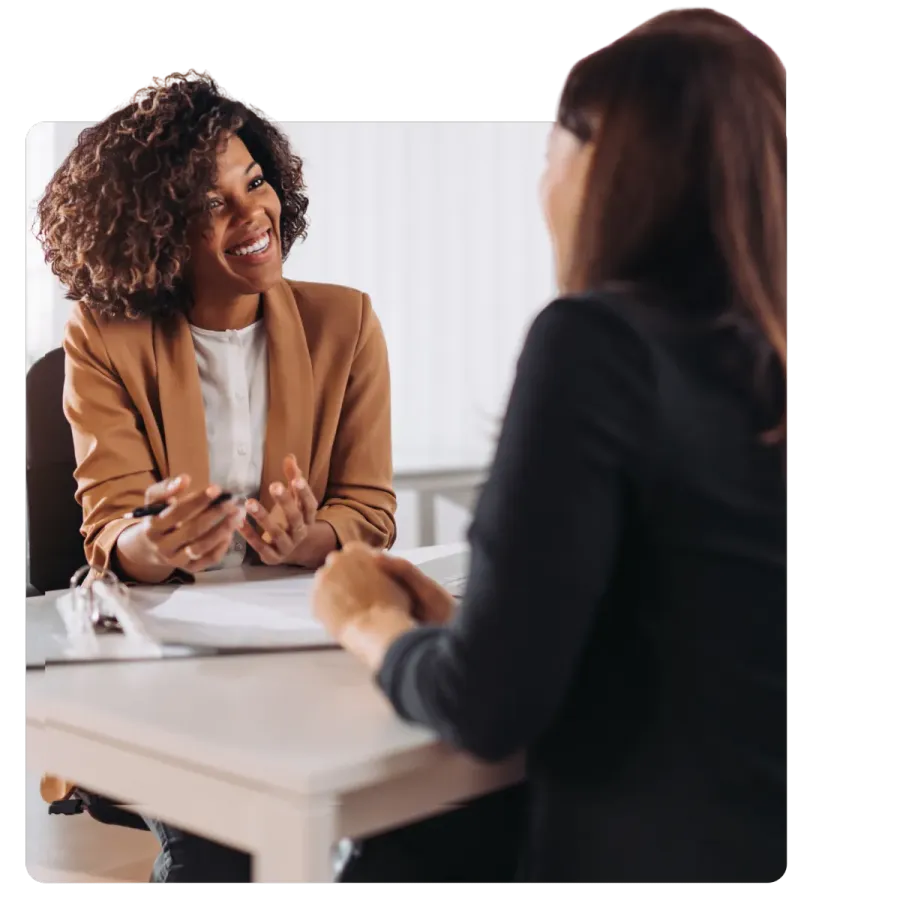 Two women in a professional setting smiling and discussing documents across a white table in an office.