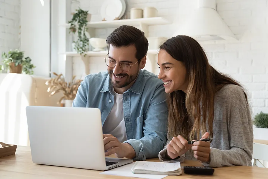Happy couple working together on laptop at bright home office desk with notebook and calculator nearby