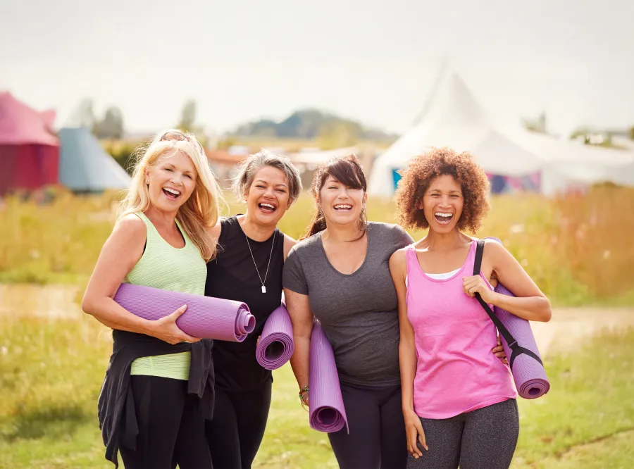 Four diverse women smiling and holding purple yoga mats outdoors in a sunny park setting.