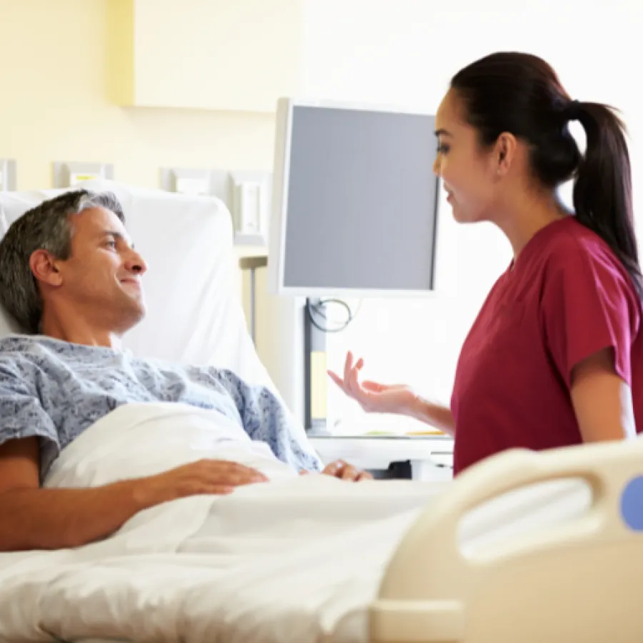 Nurse in red scrubs talking with male patient lying in hospital bed with monitor in background.