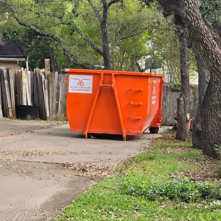Bright orange dumpster on a driveway surrounded by green trees and wooden fencing in a residential area.