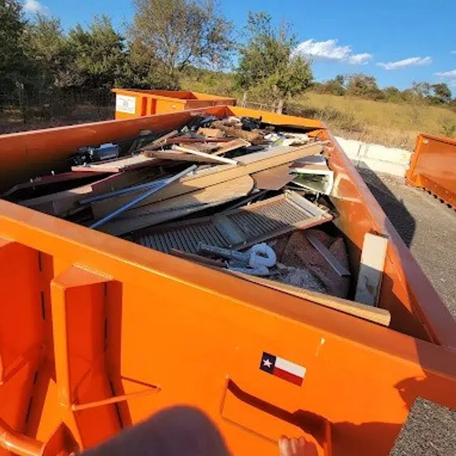 Orange dumpster filled with wooden debris and scrap materials outdoors on a sunny day with trees in background
