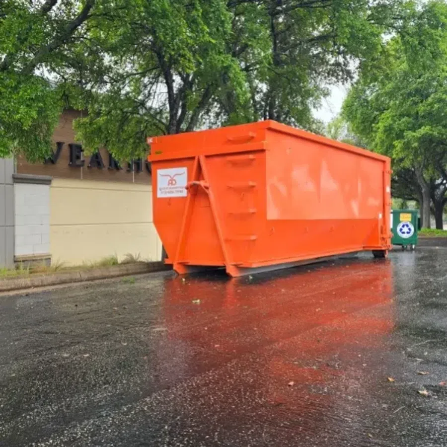 Large orange dumpster on wet pavement near trees and recycling bin outside building under cloudy sky.