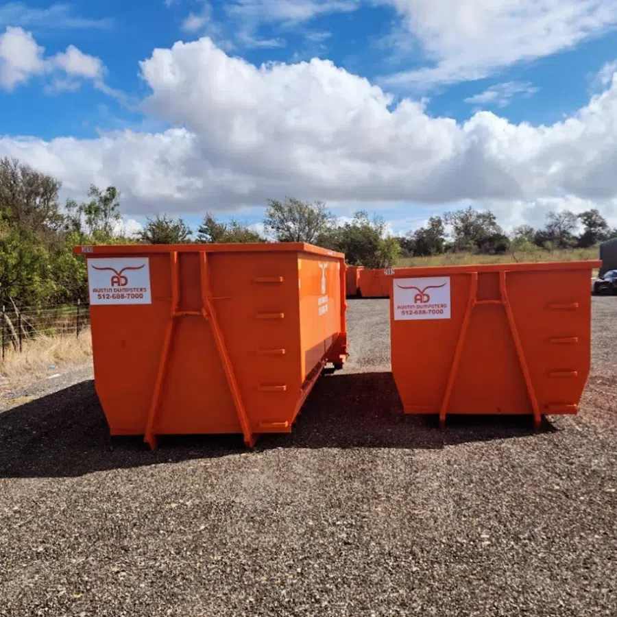 Two large orange dumpsters on gravel lot under blue sky with clouds and trees in background.