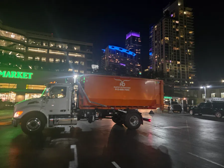 Orange Austin Dumpsters truck parked at night in a city parking lot with illuminated buildings in the background