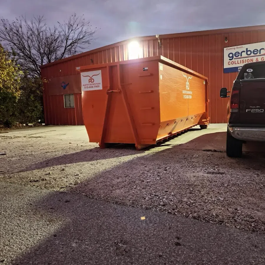 Large orange dumpster container in a gravel lot beside a pickup truck near a red building at dusk.