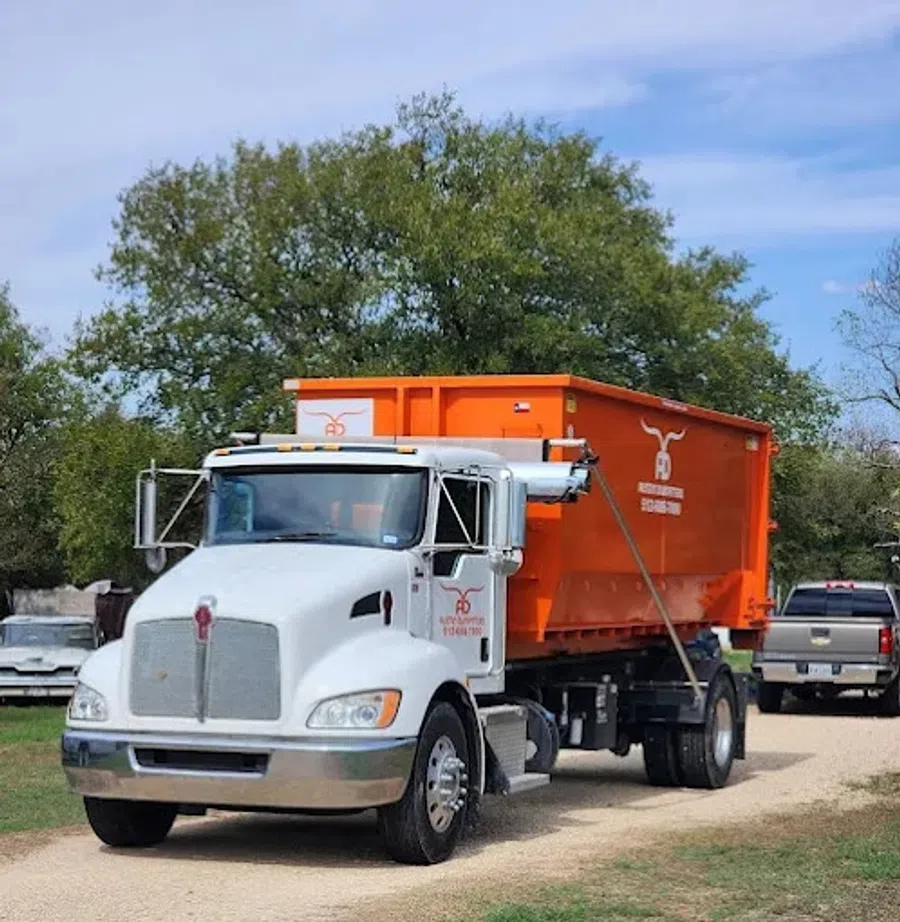 White semi truck hauling a large orange dumpster on a dirt road with trees and vehicles in the background