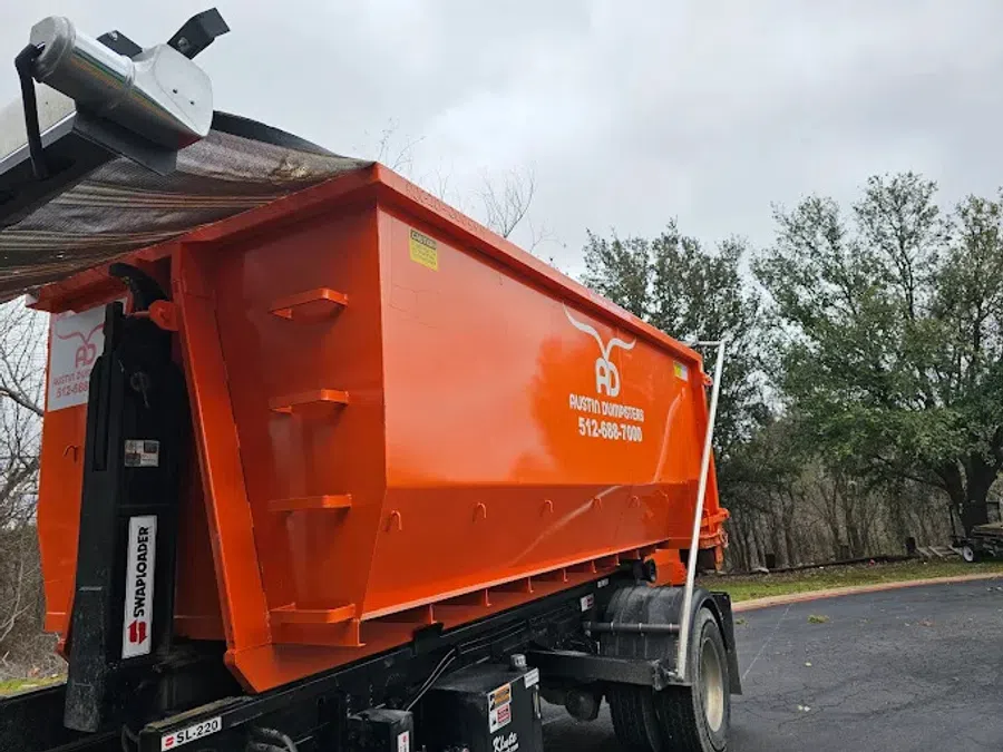 Large orange dumpster trailer with Austin Dumpsters branding parked on a driveway near trees and cloudy sky.