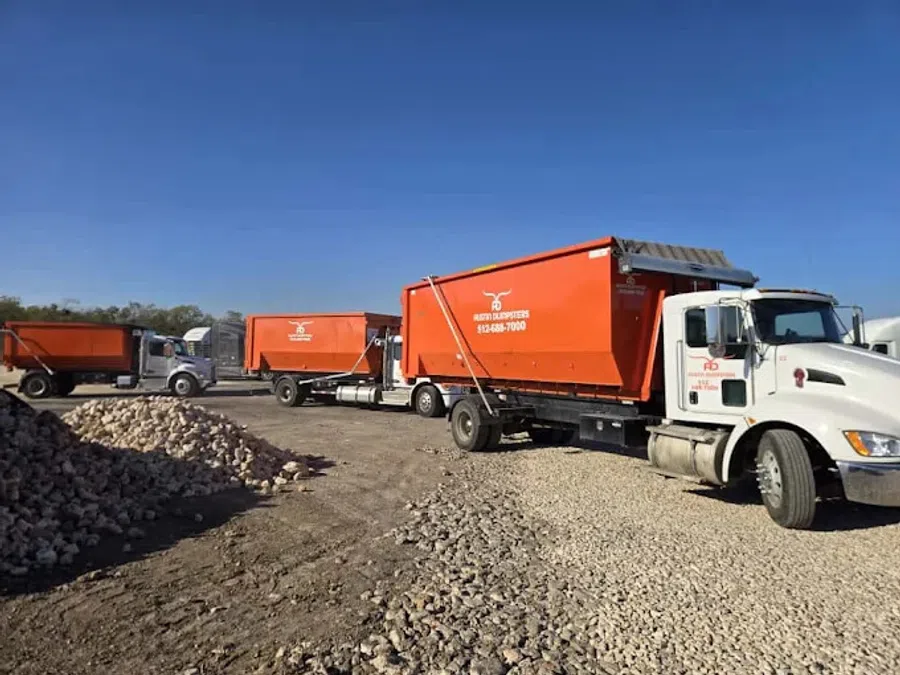 Orange dumpster trucks parked on gravel with blue sky at a construction or waste management site.