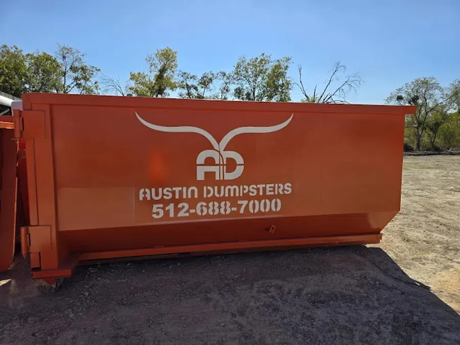 Large orange dumpster labeled Austin Dumpsters with phone number 512-688-7000 outdoors on dirt with trees
