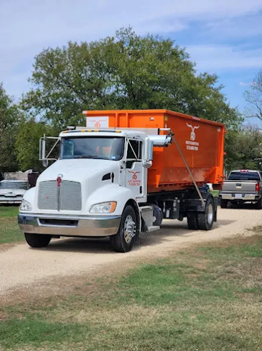 White semi-truck towing large orange container on a gravel road with green trees in the background.