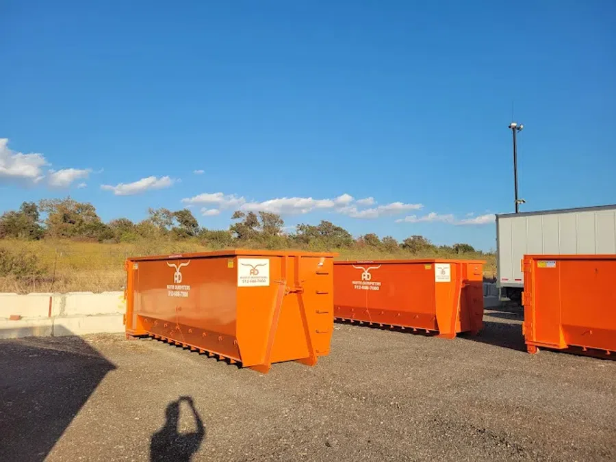 Three large orange industrial dumpsters on gravel under a clear blue sky near trees and a white building.