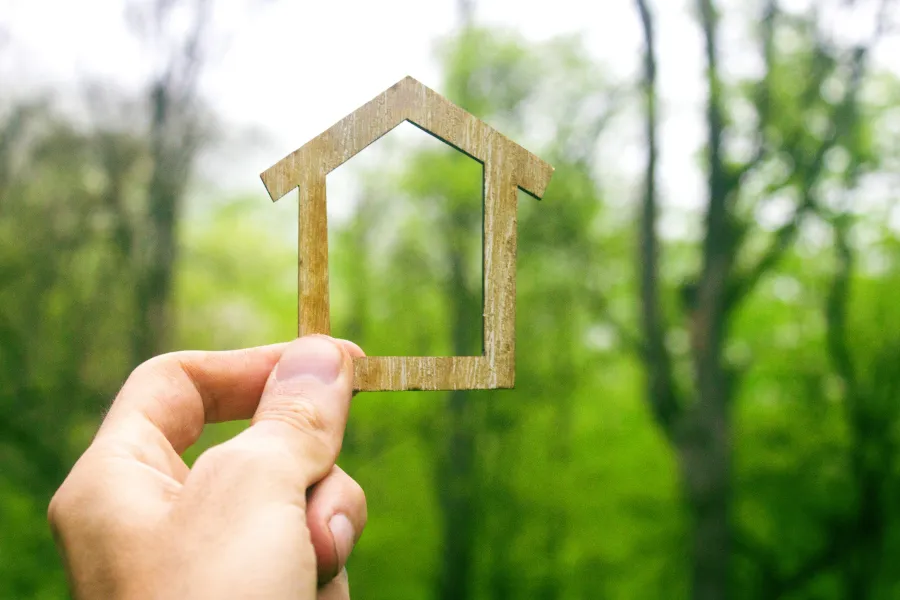 Hand holding a small wooden house frame against a blurred green forest background symbolizing home and nature.