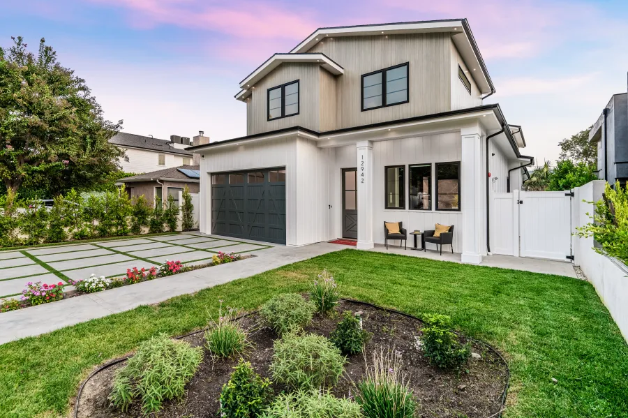 Modern two-story house with landscaped garden, driveway, and patio seating under a colorful sunset sky.