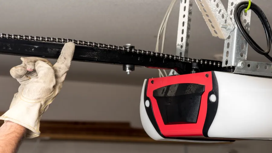 Hand in white glove adjusting garage door opener chain on ceiling-mounted motor unit with red and white casing.