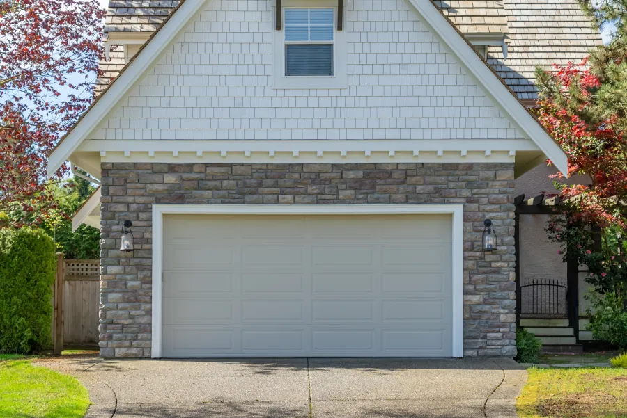 Stone and shingle garage with closed white door, framed by trees and greenery in suburban home driveway