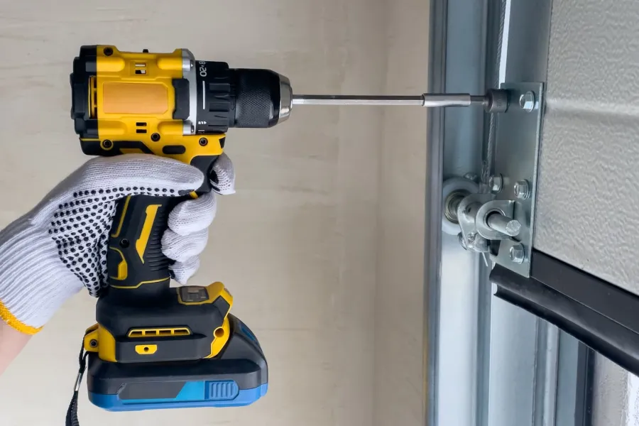 Gloved hand using a yellow cordless drill to install metal hardware on a garage door.