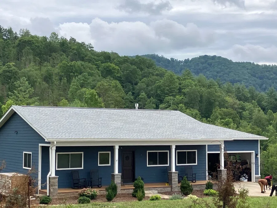 Blue single-story house with covered porch and rock columns surrounded by green forested hills under cloudy sky.