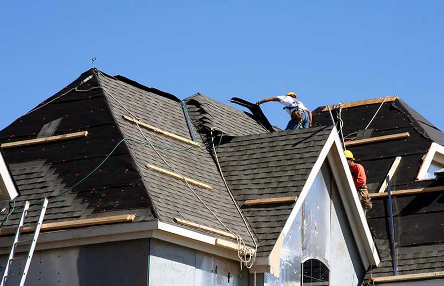 Construction workers installing black asphalt shingles on a large residential roof under a clear blue sky