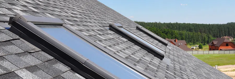 Two skylights installed on a shingled rooftop with green trees and houses in the background under a clear sky.