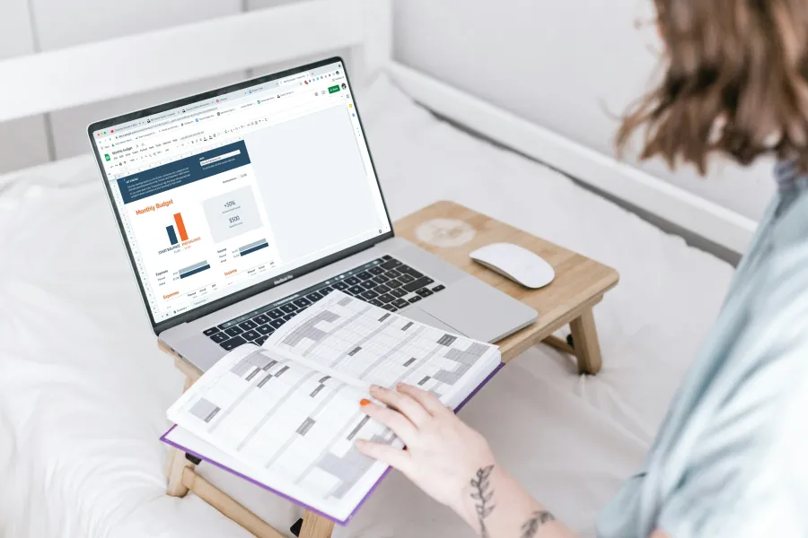 Person working on a laptop with budget charts while holding an open planner on a bed desk.