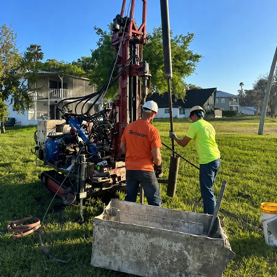 Two workers operating a drilling rig outdoors with one lifting equipment in a sunny residential area.