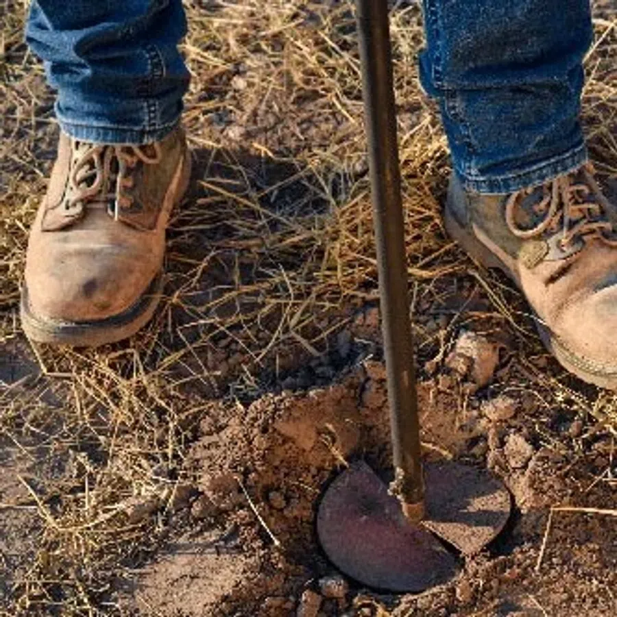 Close-up of worn boots and jeans using a hoe to dig dry soil in a field with sparse grass.