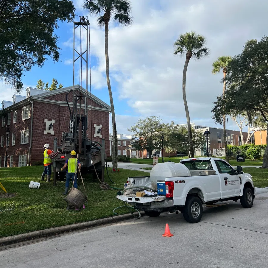 Workers in safety vests operating drilling equipment near a white utility truck on a residential street with palm trees.