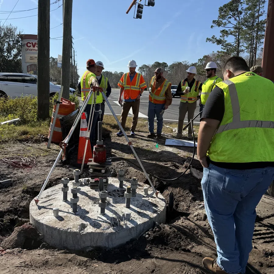 Construction workers in safety vests and helmets inspect concrete foundation with metal bolts by roadside under traffic lights.